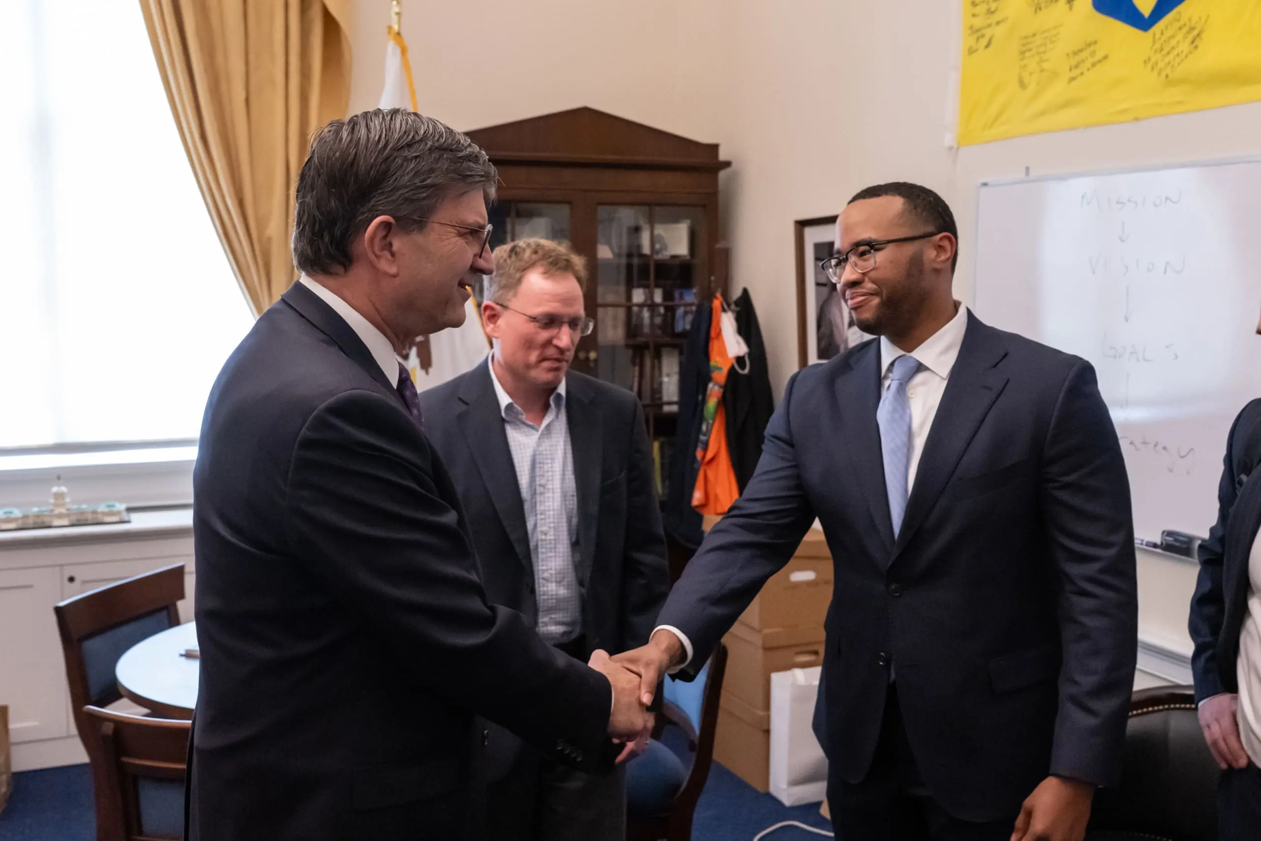 Three men in business wear stand in an office. Two of the men shake hands, and on the wall behind them is a white board with text that reads, 'Mission, Vision, Goals, Strategy'. Arrows point from one word to the next.