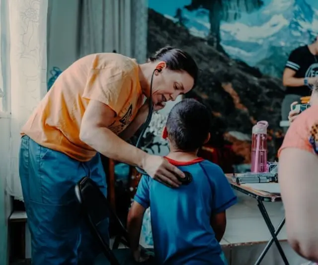 A woman leans over a young boy and holds a stethoscope to his back to listen.