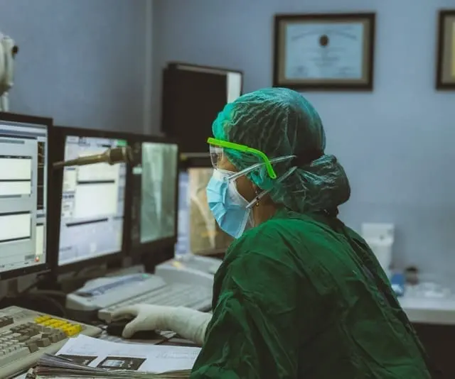 A person with a hairnet and blue mask looks at data on two computer monitors