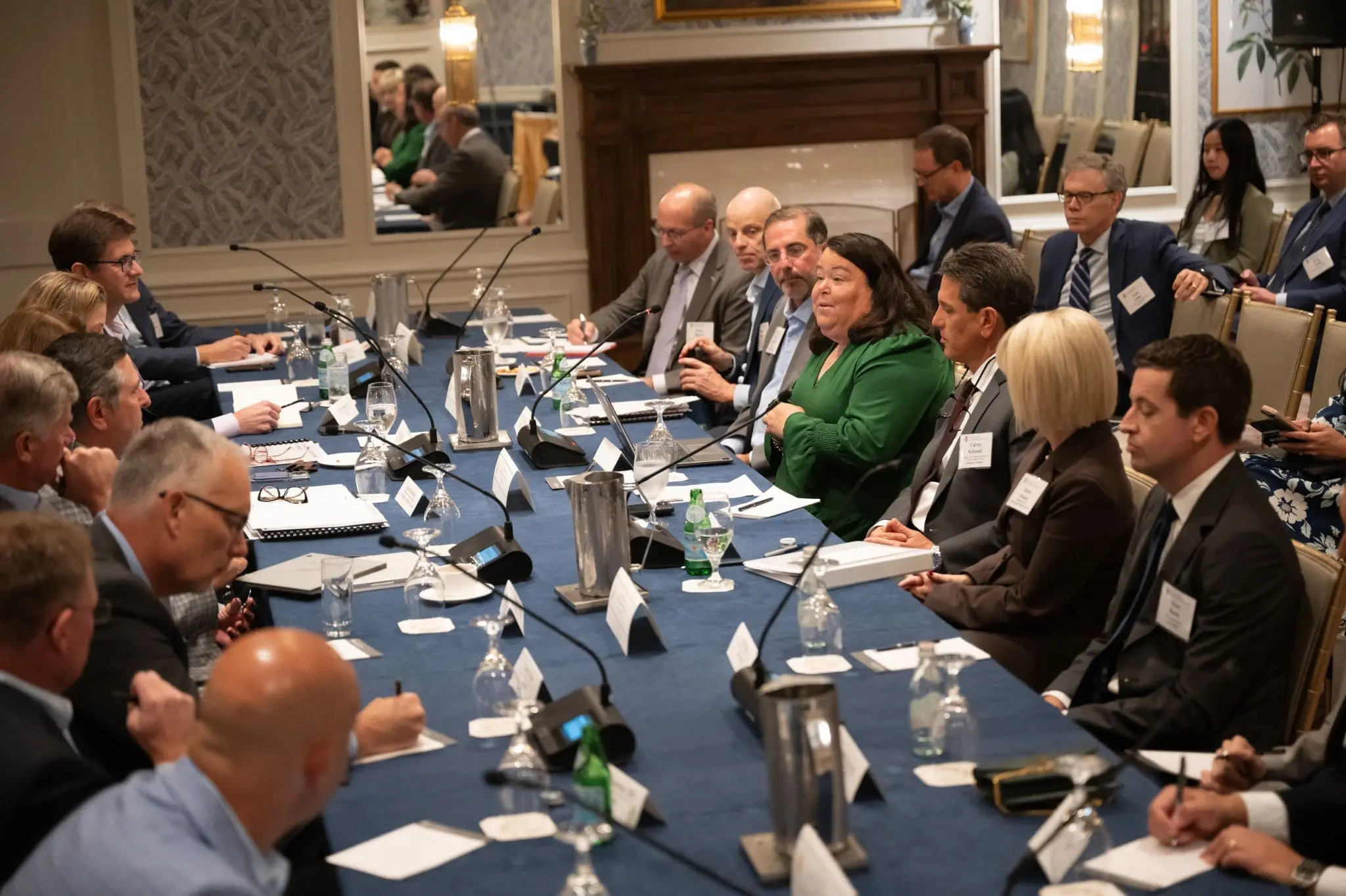 More than 20 men and women in business entire sit around a long rectangular table. Microphones, note books, laptops, and glasses sit on the table.