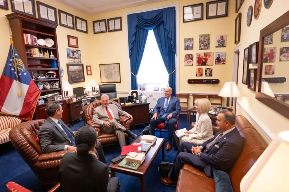 Four men and two women in business attire sit in an office around a coffee table on which is stacked several books. They sit on arm chairs, and to the left is a state flag and a bookshelf with cola bottles and sports paraphernalia on it.