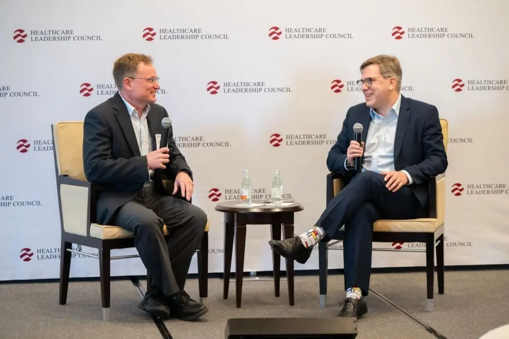 Two men sit on a stage in front of a Healthcare Leadership Council banner. They both hold microphones.