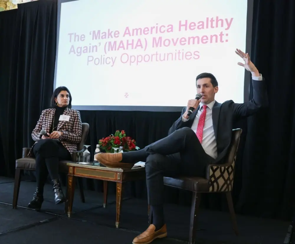 A man and a woman sit on a stage in front of a screen that reads, 'The Make America Health Again Movement: Policy opportunities.' The man sits on the right and speaks into a microphone.