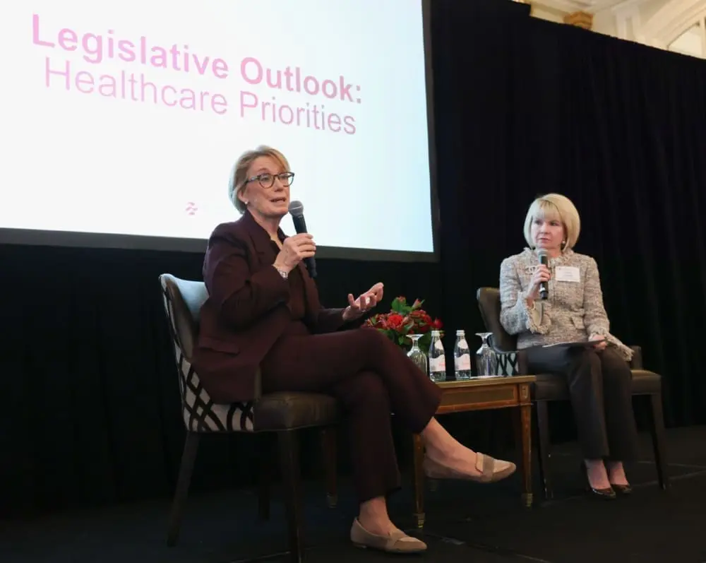 Two women with microphones sit on a stage in front of a screen that reads, 'Legislative outlook: healthcare priorities.'