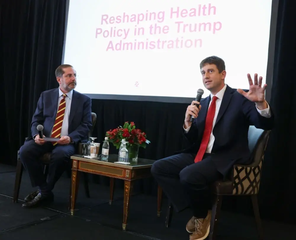 Two men sit on a stage in front of a screen that reads, 'Reshaping health: policy in the Trump administration.' The man on the right speaks into a microphone.