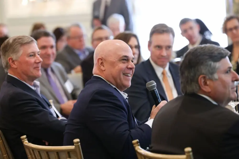 A man sits amid a group of individuals in business attire and speaks into a microphone.