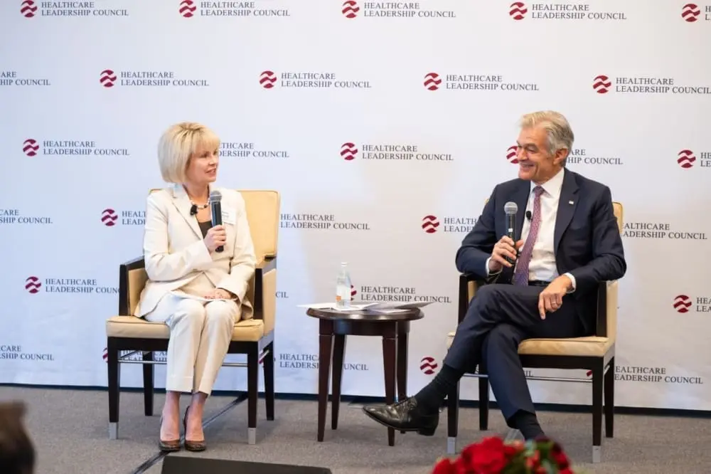 A man and a woman sit on a stage in front of a Healthcare Leadership Council banner. They both hold microphones.