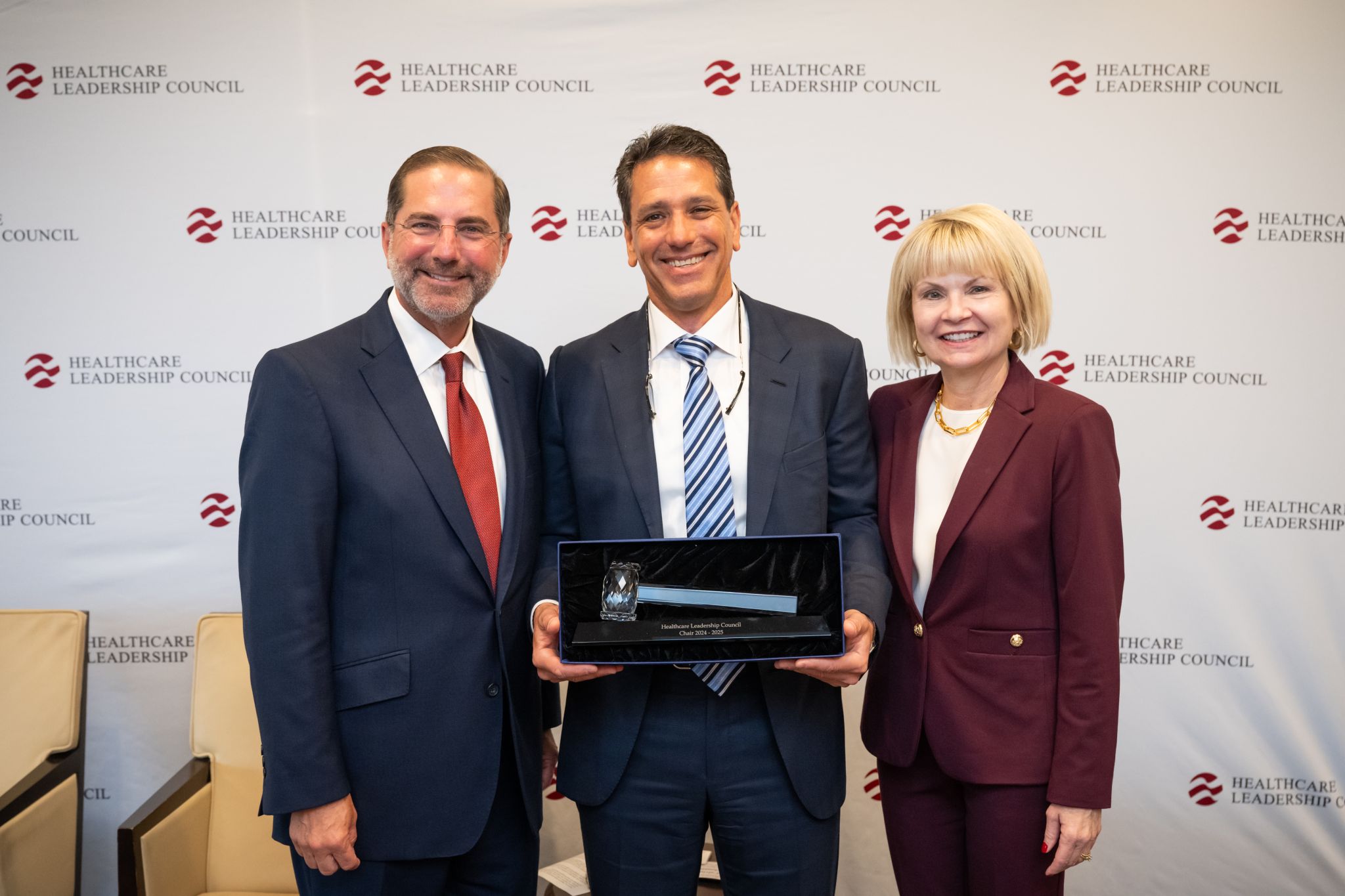 Two men and a woman who wear business suits stand in front of a wall with the Healthcare Leadership Council logo on it. The man in the middle of the group holds a crystal hammer in a velvet box, and below the hammer is text that reads 'Healthcare Leadership Council chair 2024-2025'.