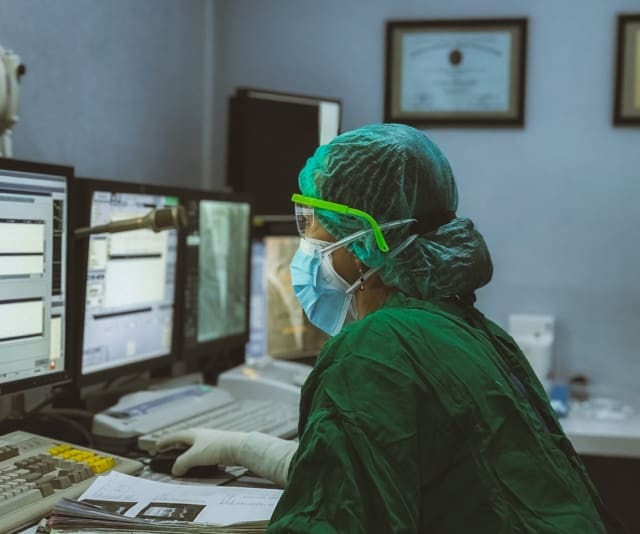 A person with a hairnet and blue mask looks at data on two computer monitors