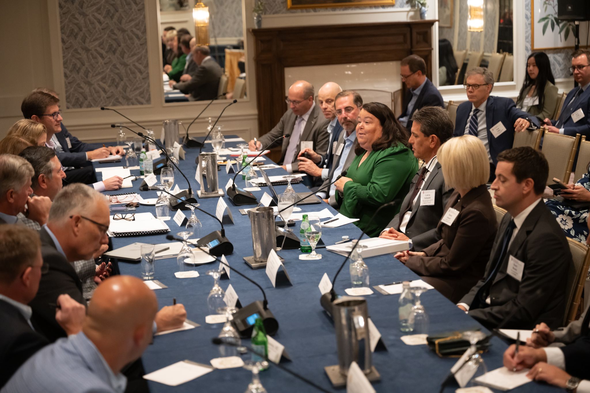 More than 20 men and women in business entire sit around a long rectangular table. Microphones, note books, laptops, and glasses sit on the table.