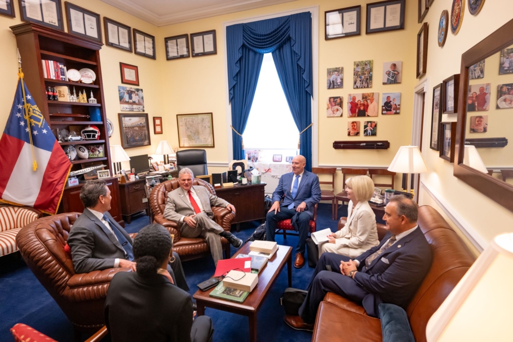 Four men and two women in business attire sit in an office around a coffee table on which is stacked several books. They sit on arm chairs, and to the left is a state flag and a bookshelf with cola bottles and sports paraphernalia on it.