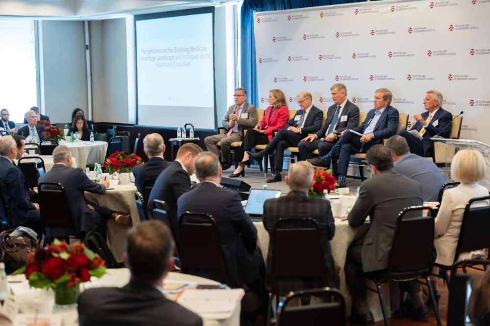 Six individuals in business attire sit on a stage in front of a room of professionals who sit around circular tables.