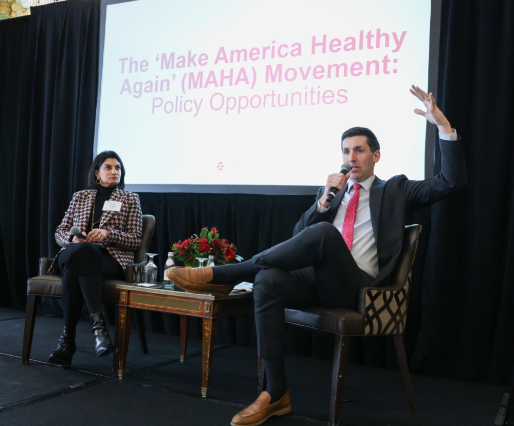 A man and a woman sit on a stage in front of a screen that reads, 'The Make America Health Again Movement: Policy opportunities.' The man sits on the right and speaks into a microphone.
