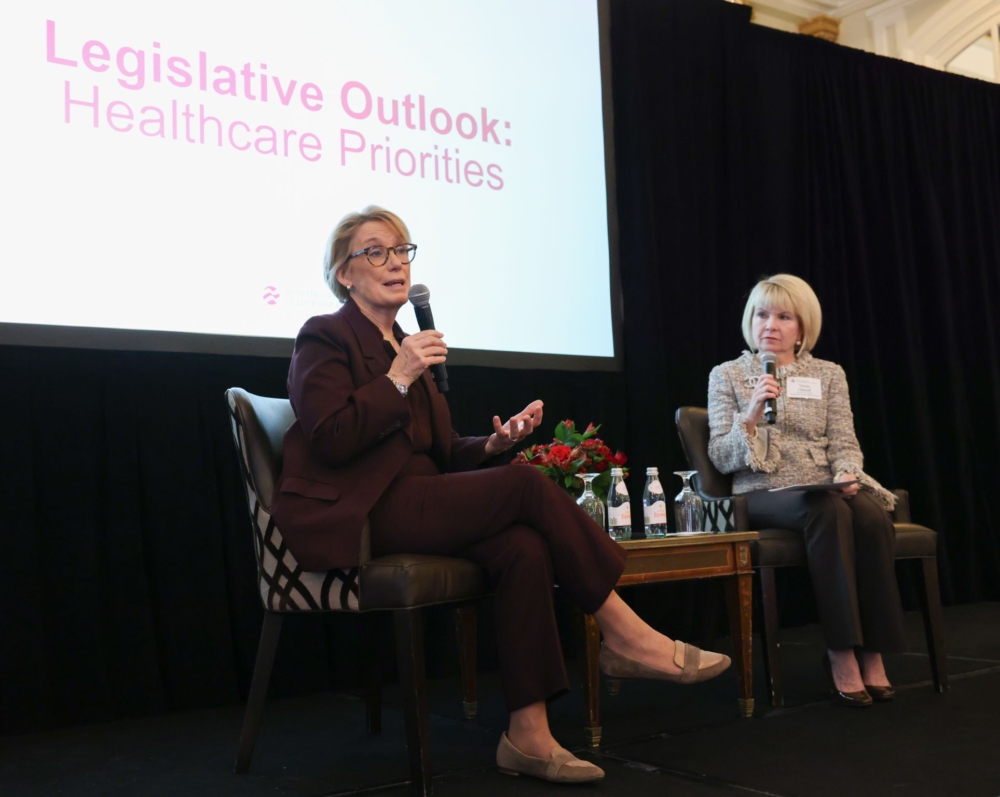 Two women with microphones sit on a stage in front of a screen that reads, 'Legislative outlook: healthcare priorities.'