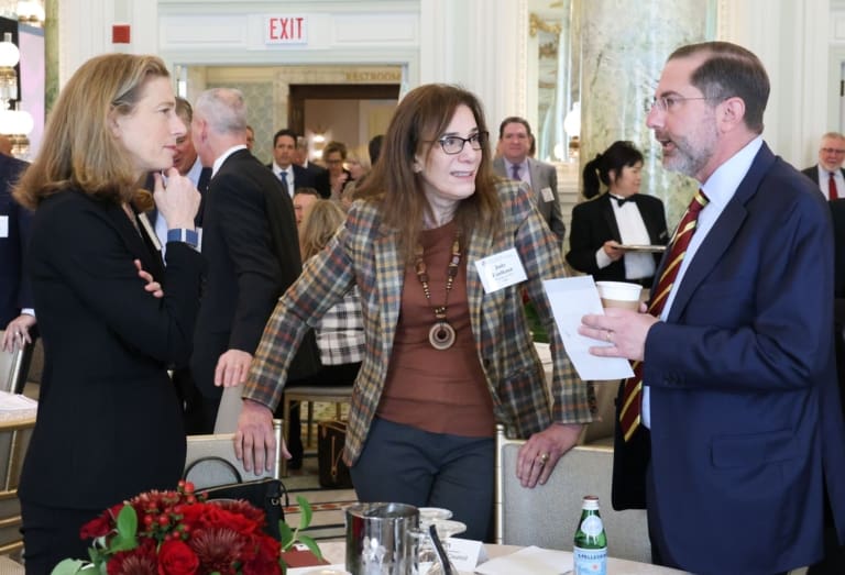 Two women in business attire look toward a man in a suit that is talking. Around them several more professionals mingle in a large room.
