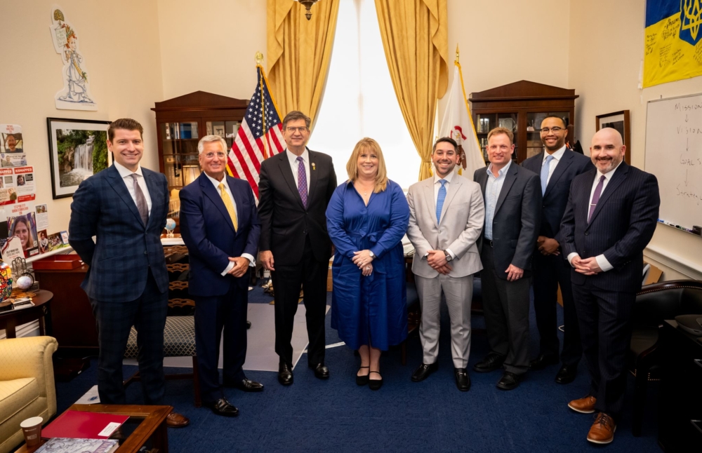 7 men and one woman stand side by side in business attire in an office. Behind them are bookcases, yellow curtains, and an American flag.