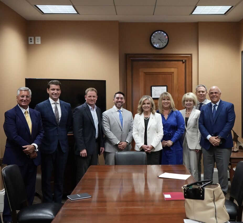 Six men and three women in business attire stand in a conference room behind a long wood table.