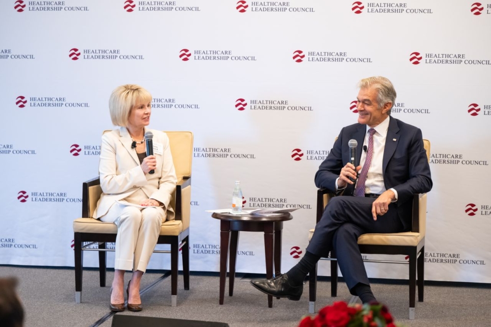 A man and a woman sit on a stage in front of a Healthcare Leadership Council banner. They both hold microphones.