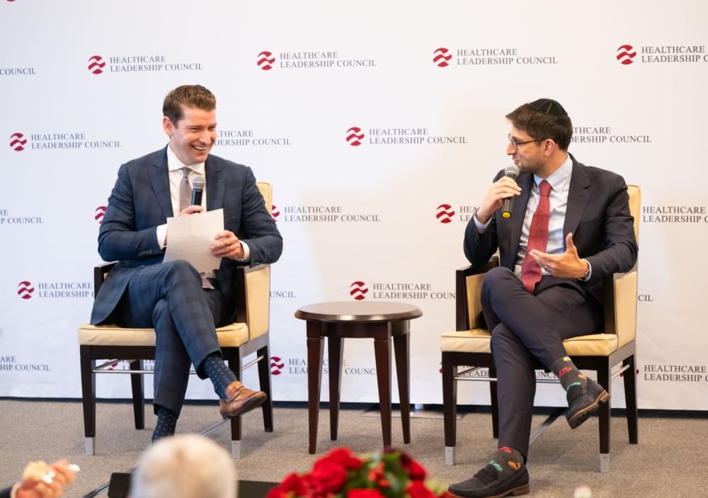 Two men sit on a stage in front of a Healthcare Leadership Council banner. They both hold microphones.
